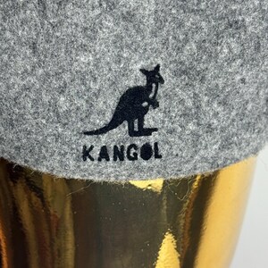 Pode incluir: Close-up de um chap&eacute;u Kangol cinzento. O chap&eacute;u apresenta um log&oacute;tipo de canguru preto e o nome da marca "KANGOL" bordado a preto. O chap&eacute;u tem um aspeto texturizado, semelhante a feltro.