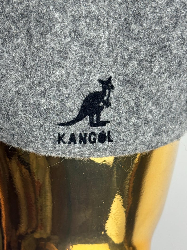 Pode incluir: Close-up de um chap&eacute;u Kangol cinzento. O chap&eacute;u apresenta um log&oacute;tipo de canguru preto e o nome da marca "KANGOL" bordado a preto. O chap&eacute;u tem um aspeto texturizado, semelhante a feltro.