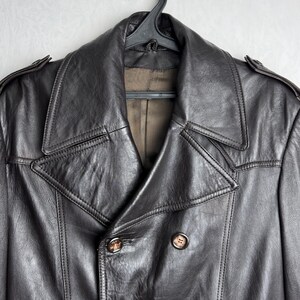 May include: A dark brown leather jacket with a double-breasted design. The jacket features a wide collar, button closures, and shoulder epaulettes. The jacket is hanging on a black hanger against a light gray background.