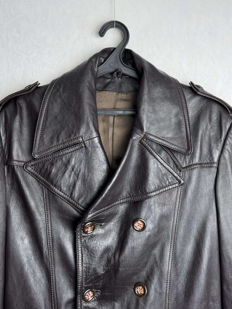 May include: A dark brown leather jacket with a double-breasted design. The jacket features a wide collar, button closures, and shoulder epaulettes. The jacket is hanging on a black hanger against a light gray background.