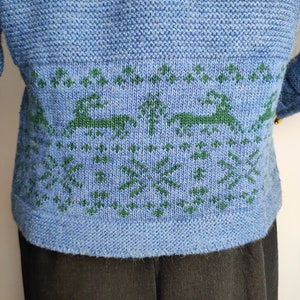 May include: A blue knitted jumper with a green knitted pattern of trees and deer. The jumper has a ribbed hem.