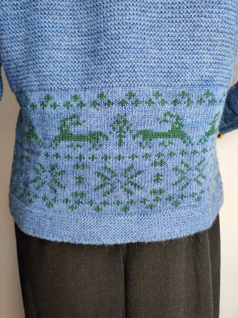 May include: A blue knitted jumper with a green knitted pattern of trees and deer. The jumper has a ribbed hem.