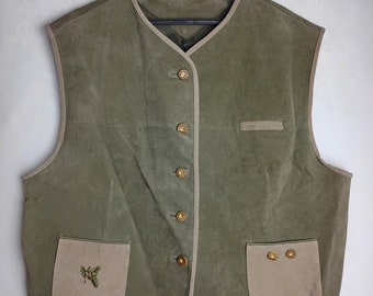 Op de afbeelding: Een groen suède vest met een crèmekleurige rand en gouden knopen. Het vest heeft twee zakken aan de voorkant, waarvan één met een klein geborduurd ontwerp.