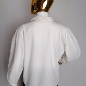 Puede incluir: Una camisa blanca de manga larga con un cuello de encaje blanco. La camisa tiene una manga ligeramente abullonada y un corte holgado.