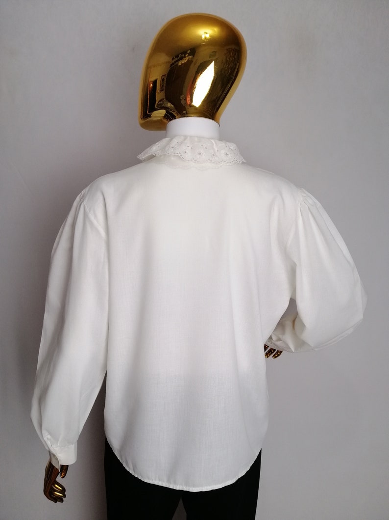Puede incluir: Una camisa blanca de manga larga con un cuello de encaje blanco. La camisa tiene una manga ligeramente abullonada y un corte holgado.