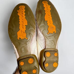 K&ouml;nnte beinhalten: Paar cremefarbene Lederschuhe mit strukturierten Oberteilen. Die Sohlen sind braun mit orangefarbenen Akzenten und dem Wort "fascine" in Orange. Die Abs&auml;tze haben orangefarbene kreisf&ouml;rmige Details. Die Schuhe werden von einer Hand gehalten.