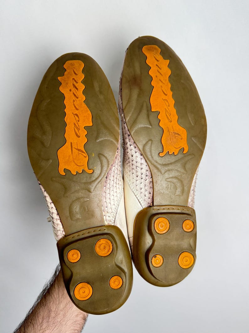 K&ouml;nnte beinhalten: Paar cremefarbene Lederschuhe mit strukturierten Oberteilen. Die Sohlen sind braun mit orangefarbenen Akzenten und dem Wort "fascine" in Orange. Die Abs&auml;tze haben orangefarbene kreisf&ouml;rmige Details. Die Schuhe werden von einer Hand gehalten.