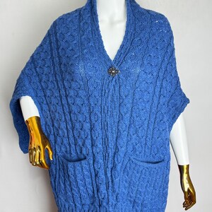 May include: A blue knitted shawl with a V-neck and two front pockets. The shawl has a cable knit pattern and is secured with a decorative brooch. The bottom edge has a fringe detail. The garment is displayed on a mannequin.