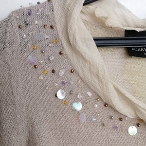 Puede incluir: Un su&eacute;ter de punto beige con una superposici&oacute;n transparente y un dise&ntilde;o decorativo de lentejuelas y cuentas en el escote. El su&eacute;ter tiene cuello en V y mangas largas. La etiqueta del su&eacute;ter dice "SCERVINO".