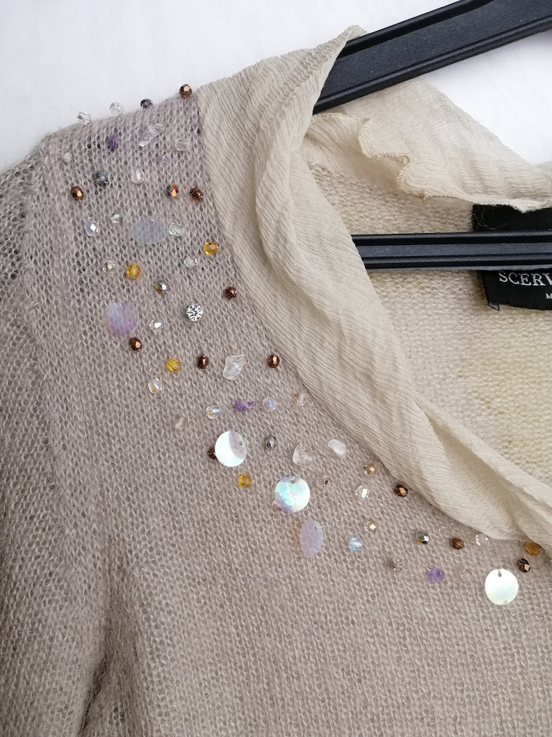 Puede incluir: Un su&eacute;ter de punto beige con una superposici&oacute;n transparente y un dise&ntilde;o decorativo de lentejuelas y cuentas en el escote. El su&eacute;ter tiene cuello en V y mangas largas. La etiqueta del su&eacute;ter dice "SCERVINO".