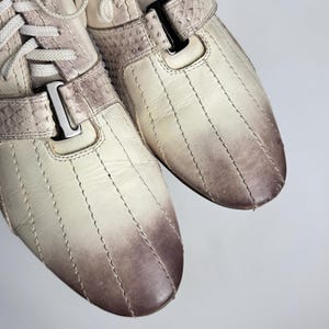 K&ouml;nnte beinhalten: Cremefarbene Sneaker mit einem Steppmuster und einem Farbverlauf von Creme zu Braun an der Spitze. Die Schuhe haben wei&szlig;e Schn&uuml;rsenkel und einen Riemen mit einer silbernen Schnalle. Das Material hat ein strukturiertes, reptilienartiges Aussehen.