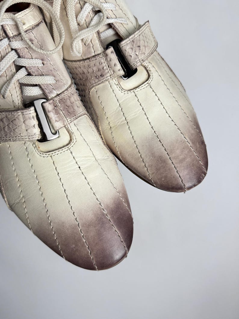 K&ouml;nnte beinhalten: Cremefarbene Sneaker mit einem Steppmuster und einem Farbverlauf von Creme zu Braun an der Spitze. Die Schuhe haben wei&szlig;e Schn&uuml;rsenkel und einen Riemen mit einer silbernen Schnalle. Das Material hat ein strukturiertes, reptilienartiges Aussehen.