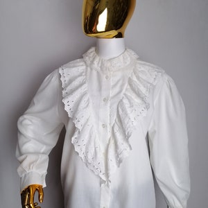 Puede incluir: Blusa blanca de manga larga con un gran cuello de encaje blanco con volantes. La blusa tiene un cierre de botones en la parte delantera y una manga ligeramente abullonada.