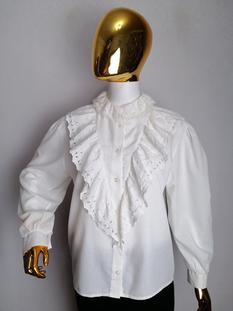 Puede incluir: Blusa blanca de manga larga con un gran cuello de encaje blanco con volantes. La blusa tiene un cierre de botones en la parte delantera y una manga ligeramente abullonada.