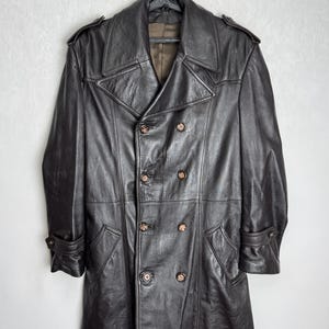 May include: A dark brown leather trench coat with a double-breasted design. The coat features a notched collar, shoulder epaulets, and multiple buttons. The coat is hanging on a black hanger.