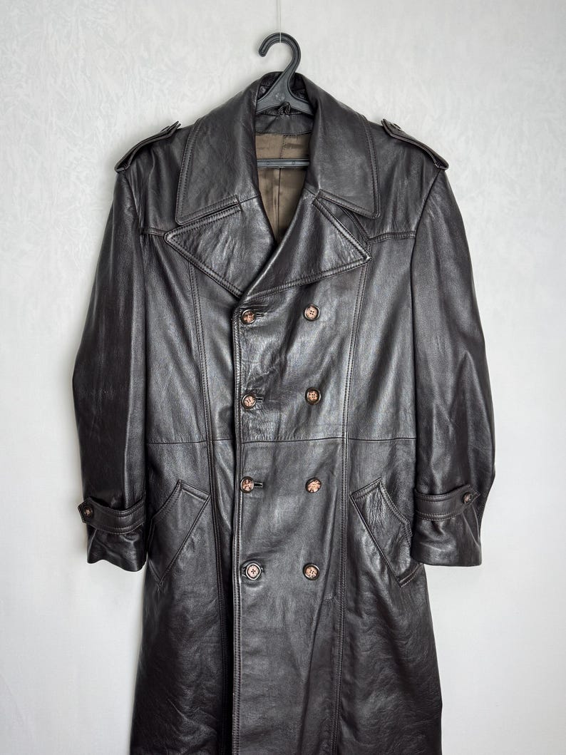 May include: A dark brown leather trench coat with a double-breasted design. The coat features a notched collar, shoulder epaulets, and multiple buttons. The coat is hanging on a black hanger.