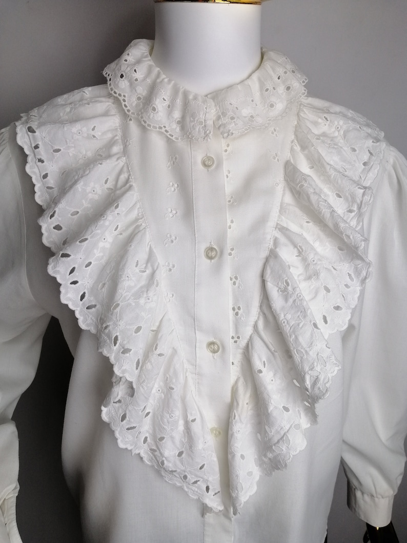 Puede incluir: Blusa blanca de algod&oacute;n con un cuello grande con volantes y detalles de ojal. El cuello est&aacute; en capas y tiene un borde festoneado.