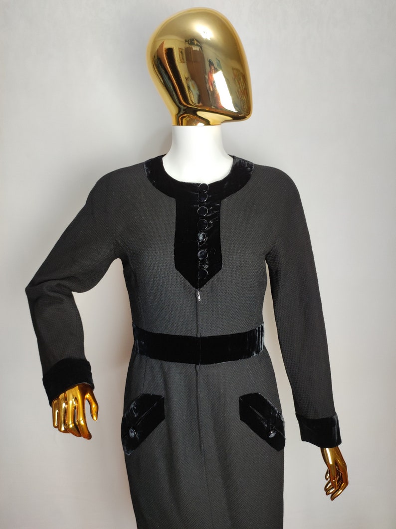 K&ouml;nnte beinhalten: Ein schwarzes Kleid mit Samtbesatz und Kn&ouml;pfen. Das Kleid hat eine figurbetonte Silhouette und einen runden Ausschnitt.