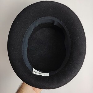 May include: A black felt hat with a rounded crown and a dark ribbon band inside the brim. The hat has a label that reads "favorite" in a script font. The hat is held up by a person's hand.