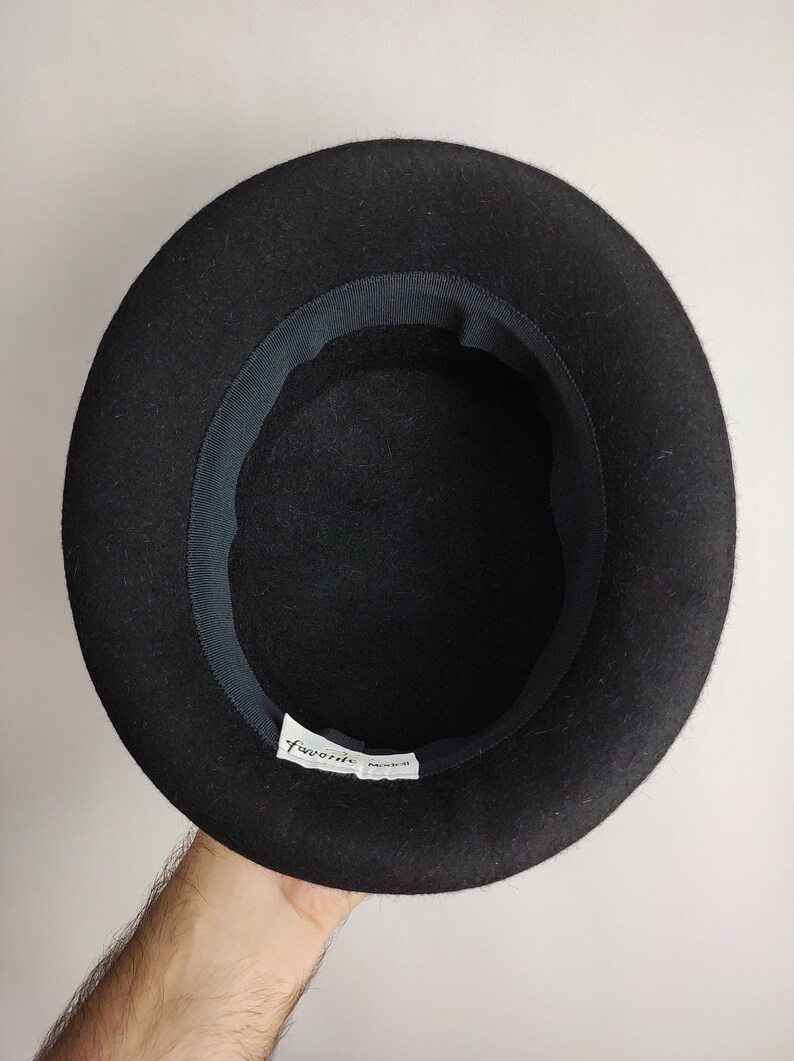 May include: A black felt hat with a rounded crown and a dark ribbon band inside the brim. The hat has a label that reads "favorite" in a script font. The hat is held up by a person's hand.