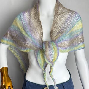 May include: A lightweight, open-knit shawl featuring a variegated color palette of soft yellow, light blue, lavender, and beige. The shawl is tied in a knot at the center front, with two loose, twisted ends hanging down, showcasing a delicate, airy texture.