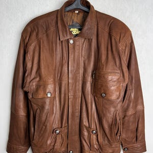 May include: Brown leather jacket with multiple pockets and button closures. The jacket has a collared neckline and a zipper pocket on the chest. The brand name "JAVAL CON" is visible on the collar. The jacket is hanging on a black hanger.