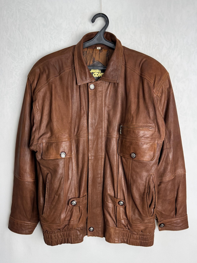 May include: Brown leather jacket with multiple pockets and button closures. The jacket has a collared neckline and a zipper pocket on the chest. The brand name "JAVAL CON" is visible on the collar. The jacket is hanging on a black hanger.