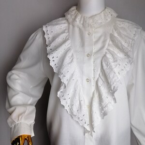 Puede incluir: Blusa blanca de manga larga con un gran cuello de volantes y ribete de encaje de ojal. La blusa tiene un cierre de botones en la parte delantera y mangas largas con pu&ntilde;os fruncidos.