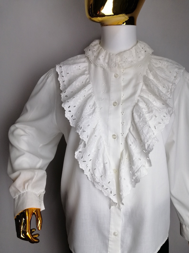Puede incluir: Blusa blanca de manga larga con un gran cuello de volantes y ribete de encaje de ojal. La blusa tiene un cierre de botones en la parte delantera y mangas largas con pu&ntilde;os fruncidos.