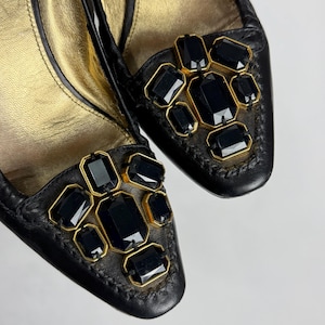 Puede incluir: Un par de bailarinas de cuero negro con interior dorado y adornos. Cada zapato presenta un grupo de gemas negras rectangulares y cuadradas engastadas en marcos dorados. Los zapatos tienen un dise&ntilde;o cosido alrededor de la puntera.