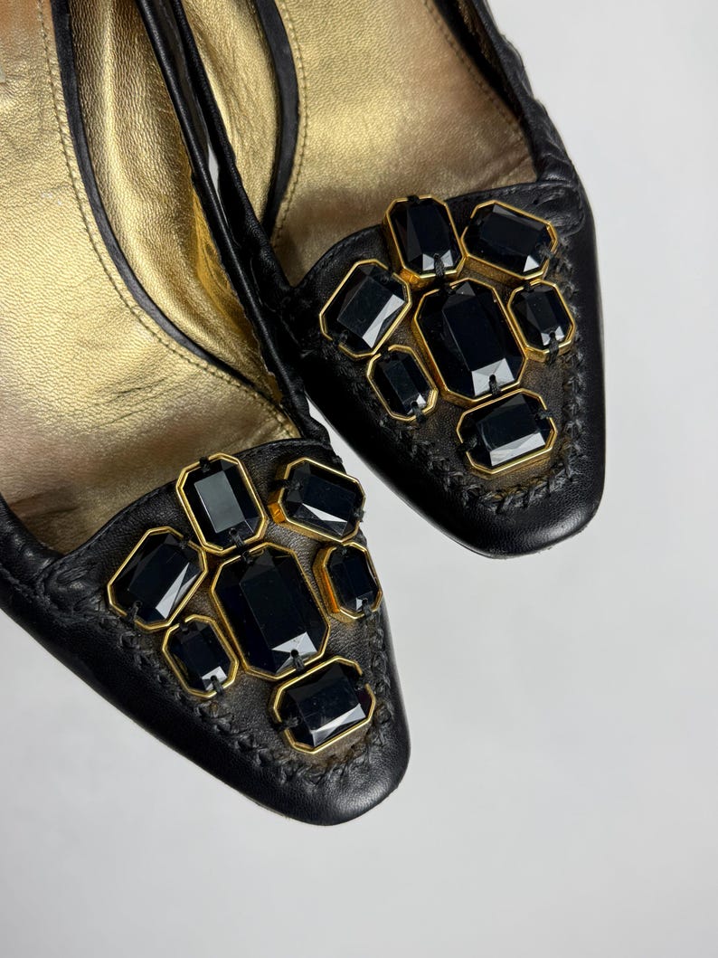 Puede incluir: Un par de bailarinas de cuero negro con interior dorado y adornos. Cada zapato presenta un grupo de gemas negras rectangulares y cuadradas engastadas en marcos dorados. Los zapatos tienen un dise&ntilde;o cosido alrededor de la puntera.