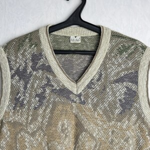 May include: A vintage V-neck sweater vest with a patterned design in shades of beige, gray, green, and red. The vest is made of a knit material and has a ribbed trim around the neckline and armholes. The vest is hanging on a black hanger.