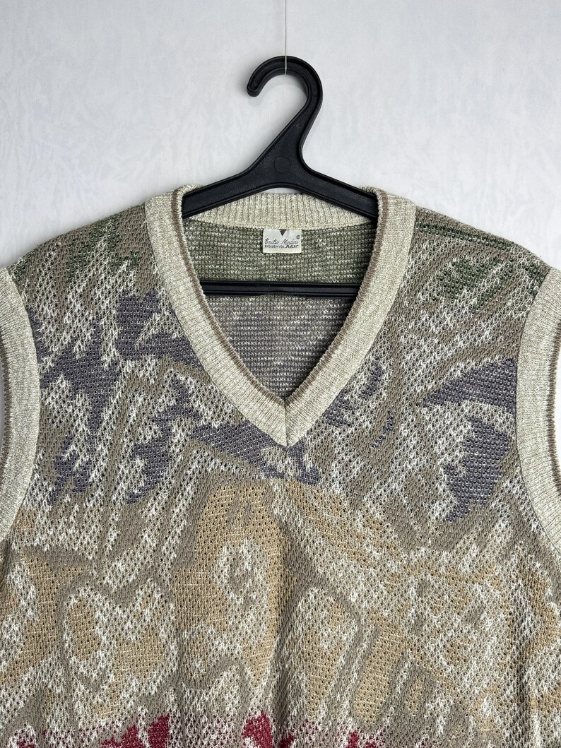 May include: A vintage V-neck sweater vest with a patterned design in shades of beige, gray, green, and red. The vest is made of a knit material and has a ribbed trim around the neckline and armholes. The vest is hanging on a black hanger.