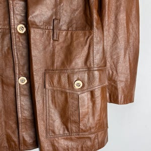 May include: A brown leather jacket with multiple pockets and gold-colored buttons. The jacket has a classic design with a button-up front and a tailored fit. The leather appears to be well-worn, giving it a vintage look.