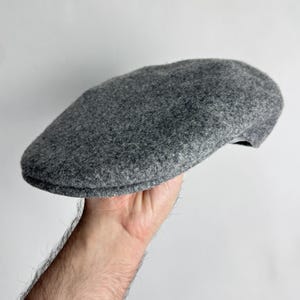 Pode incluir: Bon&eacute; plano cinzento de l&atilde;. O bon&eacute; &eacute; de cor cinza-m&eacute;dio liso e tem uma coroa arredondada e uma viseira pequena e r&iacute;gida. O bon&eacute; &eacute; segurado por uma m&atilde;o sobre um fundo branco. Este estilo cl&aacute;ssico &eacute; adequado para v&aacute;rias ocasi&otilde;es.