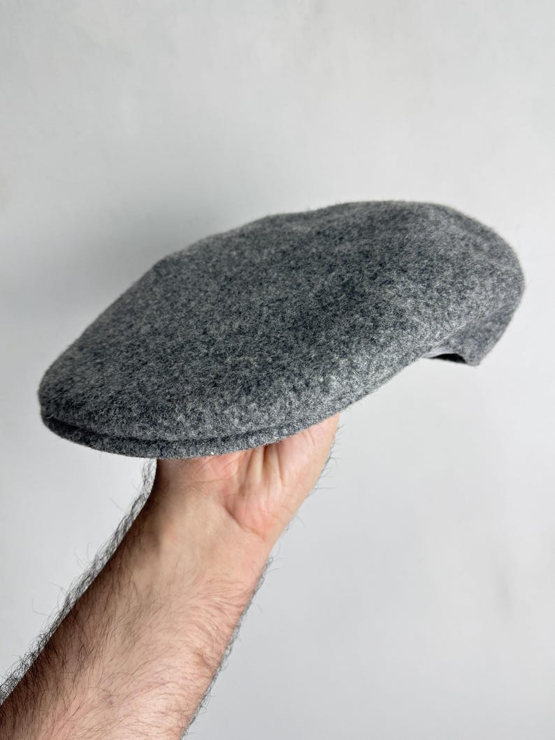 Pode incluir: Bon&eacute; plano cinzento de l&atilde;. O bon&eacute; &eacute; de cor cinza-m&eacute;dio liso e tem uma coroa arredondada e uma viseira pequena e r&iacute;gida. O bon&eacute; &eacute; segurado por uma m&atilde;o sobre um fundo branco. Este estilo cl&aacute;ssico &eacute; adequado para v&aacute;rias ocasi&otilde;es.