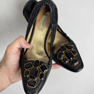Puede incluir: Bailarinas de cuero negro con detalles dorados y grandes gemas oscuras. Los zapatos presentan un dise&ntilde;o cosido en los bordes y un interior dorado. La marca "PRADA" es visible en el interior.