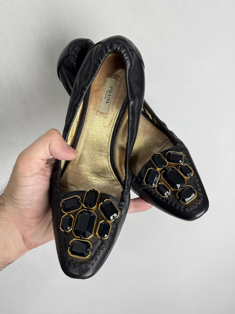 Puede incluir: Bailarinas de cuero negro con detalles dorados y grandes gemas oscuras. Los zapatos presentan un dise&ntilde;o cosido en los bordes y un interior dorado. La marca "PRADA" es visible en el interior.