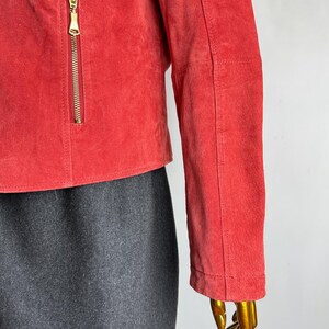 Puede incluir: Chaqueta corta de ante rojo con cremallera dorada y corte entallado. Se ve la manga de la chaqueta, junto con una parte de una falda gris. La chaqueta se exhibe en un maniqu&iacute;.