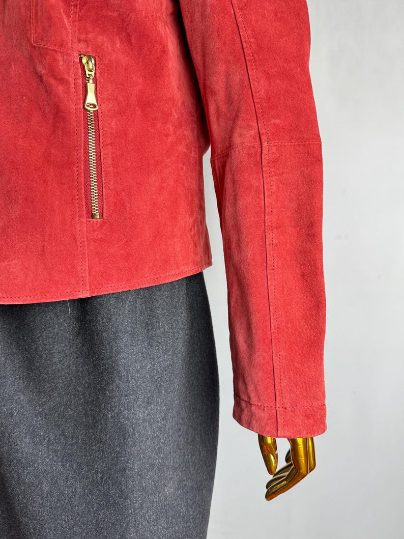 Puede incluir: Chaqueta corta de ante rojo con cremallera dorada y corte entallado. Se ve la manga de la chaqueta, junto con una parte de una falda gris. La chaqueta se exhibe en un maniqu&iacute;.