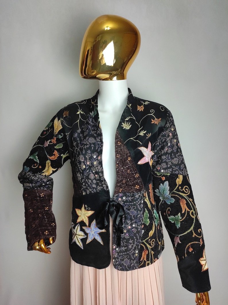 May include: A black velvet jacket with a patchwork design featuring embroidered floral motifs and sequins. The jacket has a tie closure at the waist.