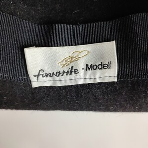 May include: Close-up of a black hat with a black grosgrain ribbon band. A white label with gold script and the words "favorite - Modell" is sewn into the hat.