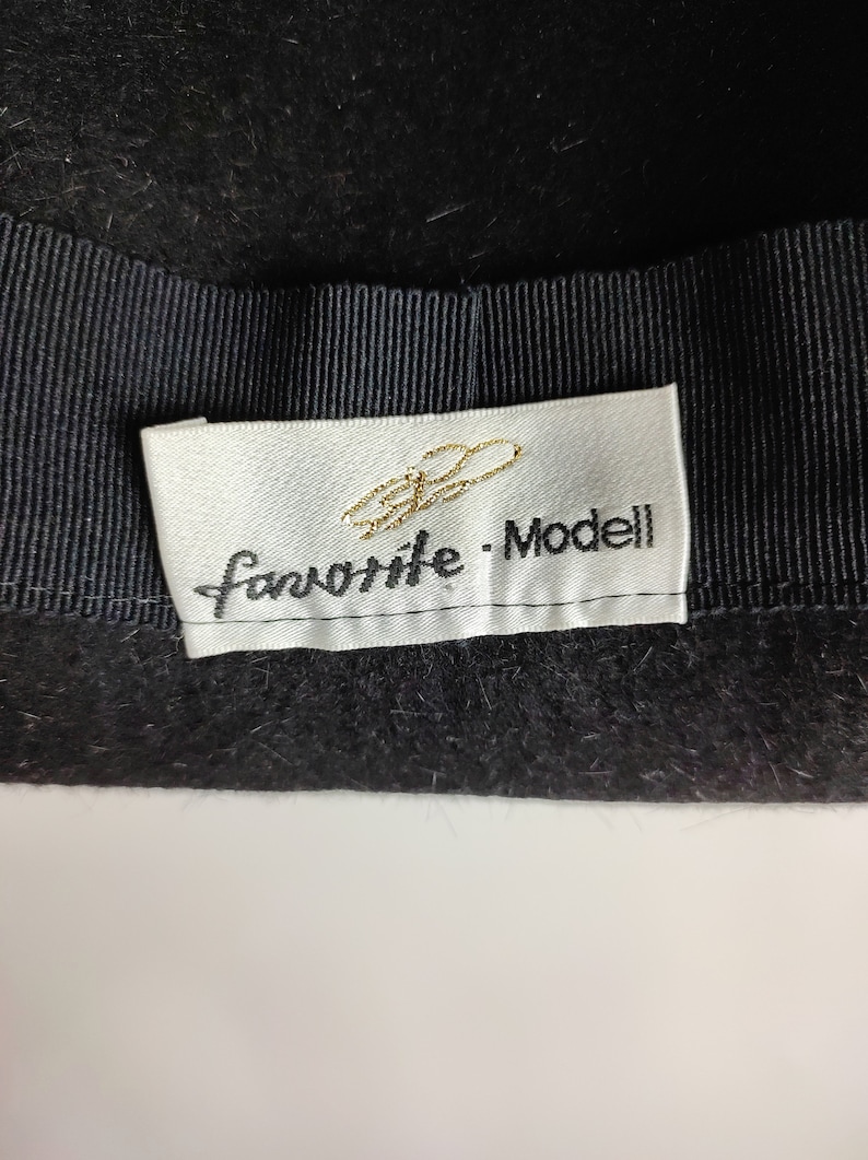 May include: Close-up of a black hat with a black grosgrain ribbon band. A white label with gold script and the words "favorite - Modell" is sewn into the hat.
