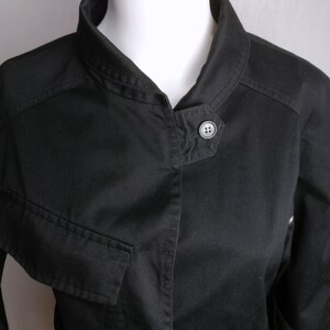 May include: A black utility jacket with a stand-up collar, a button closure, and a belt at the waist.