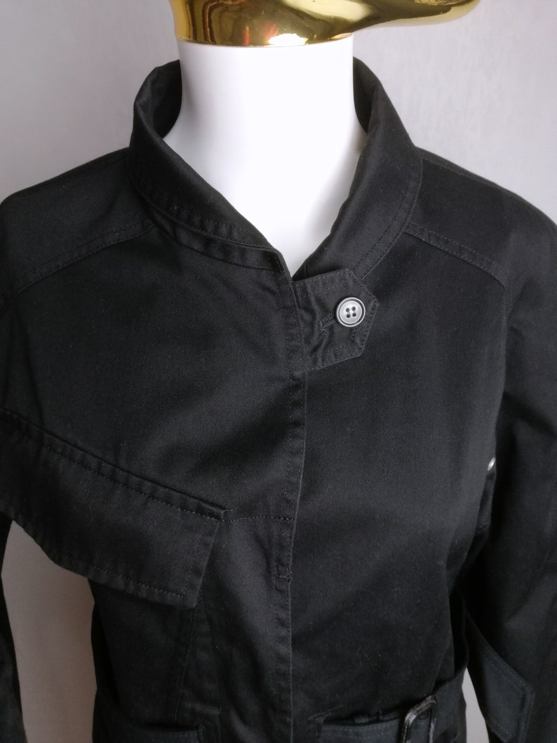 May include: A black utility jacket with a stand-up collar, a button closure, and a belt at the waist.