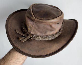 Puede incluir: Un sombrero vaquero de cuero marrón con una banda de cuero trenzada y un ala ancha.