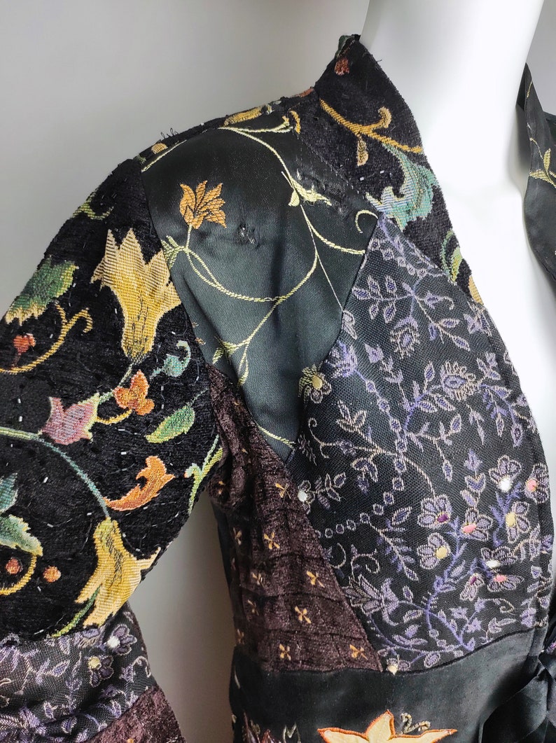 May include: A black velvet jacket with a patchwork design featuring floral patterns in shades of gold, purple, and brown. The jacket has a long sleeve and a V-neck.