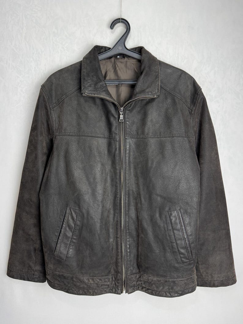 Puede incluir: Chaqueta de cuero marr&oacute;n con cremallera y dos bolsillos delanteros. La chaqueta tiene cuello y est&aacute; colgada de una percha negra. El cuero parece desgastado, d&aacute;ndole un aspecto vintage. La chaqueta es de talla mediana.