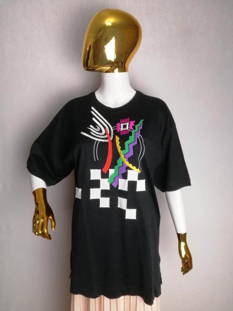May include: Black t-shirt with a colourful abstract design featuring geometric shapes, lines, and a chequered pattern.