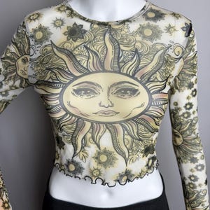 May include: A white mesh long-sleeved crop top with a black and yellow sun graphic. The sun has a face and is surrounded by floral and paisley designs.